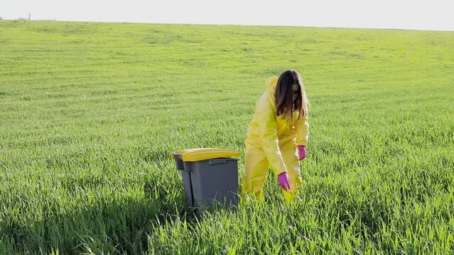 A woman in a yellow protective suit stands in the middle of a green field and holds a plastic bottle in her hands to throw in the tank