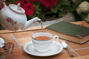 Elegance, romance and- someone pours tea from a porcelain teapot into a cup on a vintage table in garden against a background of flowers and an old book in summer