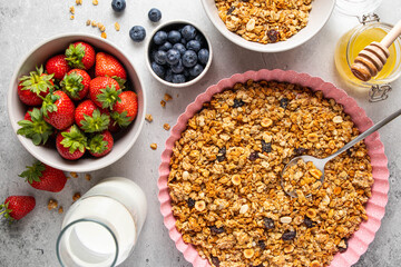 Homemade granola with raisin, seeds, hazelnut and peanut on baking sheet, milk or yogurt bottle, honey and blueberries, strawberries on grey background. Healthy breakfast ingredients. Selective focus.