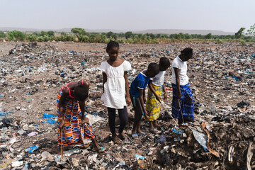 Group of children rummaging through a field covered with waste to find recyclable material to sell,...