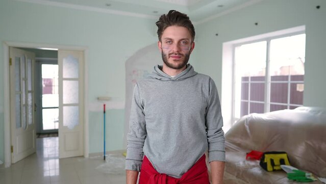 Front view cheerful man coloring face with paint looking at camera smiling. Positive handsome brunette Caucasian guy posing indoors in new house painting walls