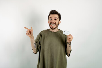 Happy casual man wearing t-shirt posing isolated over white background holding credit bank card and pointing index finger aside. Shopping and finance concept.