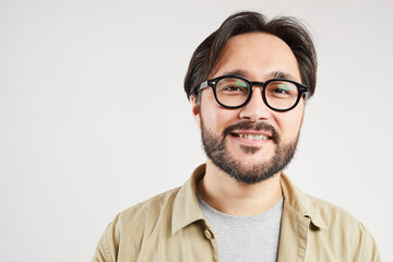 Cheerful young Asian man with beard wearing casual shirt and glasses standing against white background and looking at camera