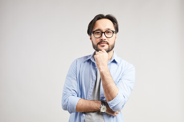 Portrait of content enterprising young Asian man with beard wearing casual shirt and eyeglasses touching chin in thoughts