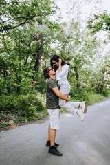 Fototapeta premium A bearded long-haired stylish man holds in his arms a brunette hippie woman in sunglasses gently hugs and smiles in nature in the park. Portrait, photograph of people in love.