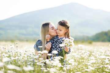 Cute mother kissing child girl daughter in meadow with blooming flowers outdoor together. Family...