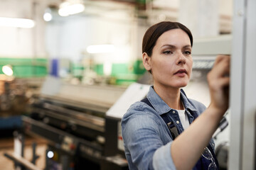 Fototapeta premium Busy focused brunette female factory engineer standing in modern industrial shop and choosing program on cnc lathe