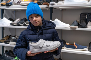 Satisfied boy in winter clothes chooses fashionable white shoes for the spring season in shoe store