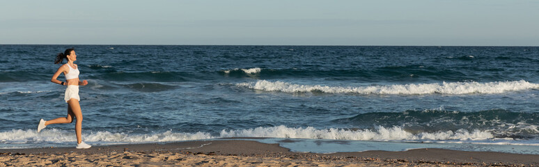 full length of young sportive woman jogging near sea in summer, banner.