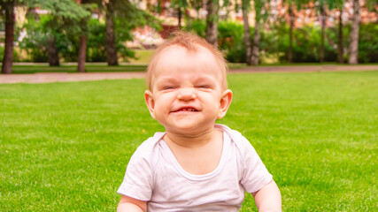 Cute laughing blonde baby girl 1 year old top having fun sitting in green grass outdoors. Childhood. Summer season. Looking at camera. Happy little child laughs nicker in meadow.