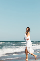 Fototapeta na wymiar full length of joyful woman in white shirt and swimsuit smiling near ocean on beach.