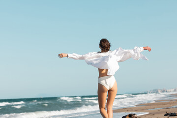 back view of young woman in white shirt and swimwear standing with outstretched hands near ocean on beach.