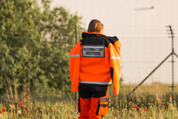 Woman a polish ambulance worker standing back in medical orange uniform with inscription Emergency...