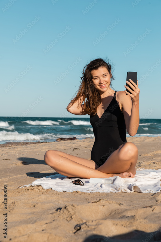 Wall mural happy young woman in swimsuit taking selfie on smartphone while sitting on beach.