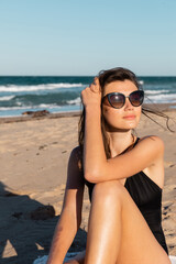 sunshine on face of young woman in black swimwear and sunglasses sitting on beach near sea.