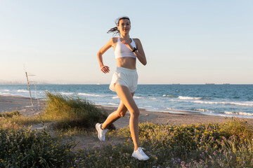 smiling young woman in shorts and wireless earphones jogging on grass near sea.