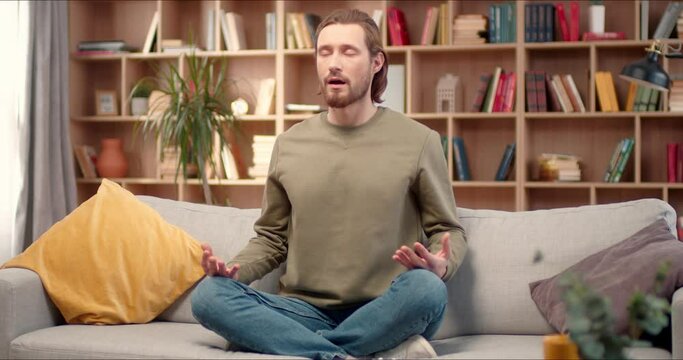 Close up portrait on young handome caucasian man sitting in lotus pose on couch. Calm young man tries to meditate in living room. Peaceful handsome white guy meditating for concentration, calmness