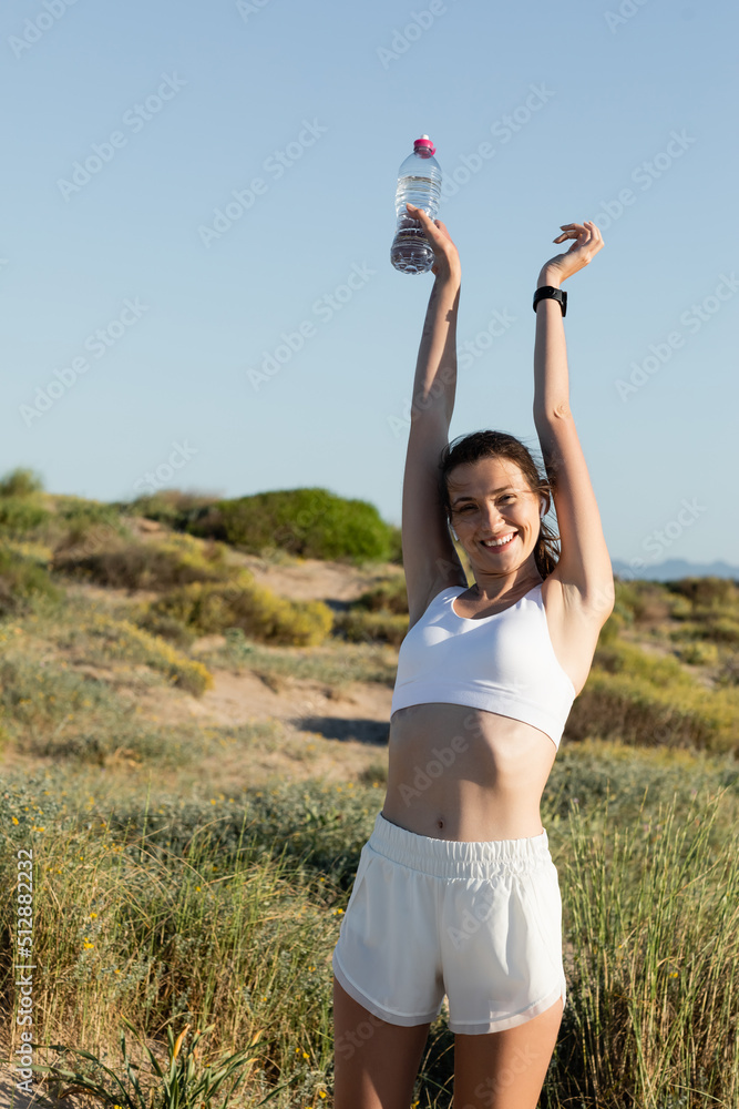 Wall mural happy sportswoman in wireless earphones holding bottle while stretching outside.