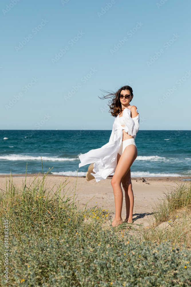 Wall mural full length of brunette young woman in sunglasses and white shirt holding straw hat on sandy beach.