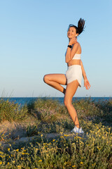 happy fit woman in sportive shorts and wireless earphone jumping near blue sea.