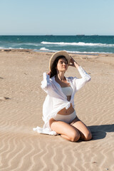 Fototapeta na wymiar young woman in white shirt and swimwear wearing straw hat and sitting on golden sand near blue ocean.
