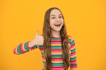 Teenager child girl 12, 13, 14 year old doing thumbs up gesture. Cheerful teenager girl showing thumbs up sign, isolated on yellow background.