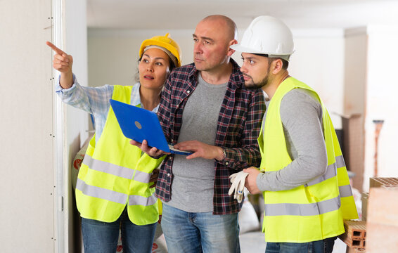 Interested adult male homeowner discussing home renovation work plan with builders while standing inside with laptop in hands..