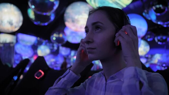 Woman wearing wireless black headphones and looking around in dark room of interactive exhibition or museum with colorful illumination. Futuristic, entertainment, immersive concept