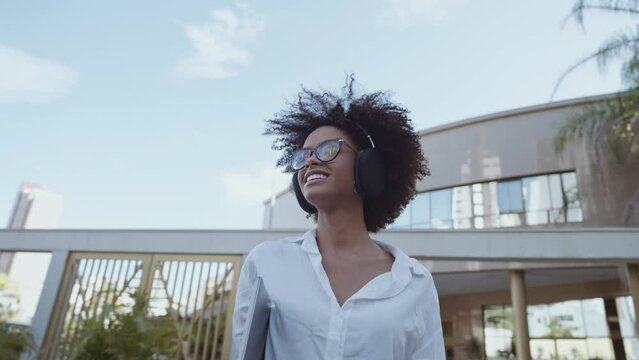 Low angle view of smiling Latin businesswoman holding laptop and eyeglasses near blurred building. Cinematic 4K