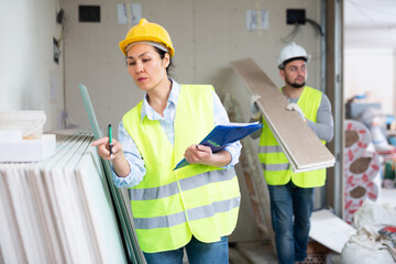 Strict focused asian female building inspector wearing yellow safety vest and hard hat standing with papers at construction site indoors, taking inventory of materials