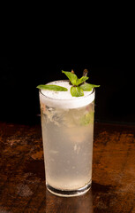 refreshing long drink with foam, mint on wooden table on black background angle view
