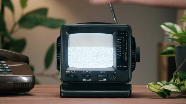 Knocking on small old television with grey interference screen on home background. Close-up of vintage tv on table with retro phone and plants, nostalgia. 