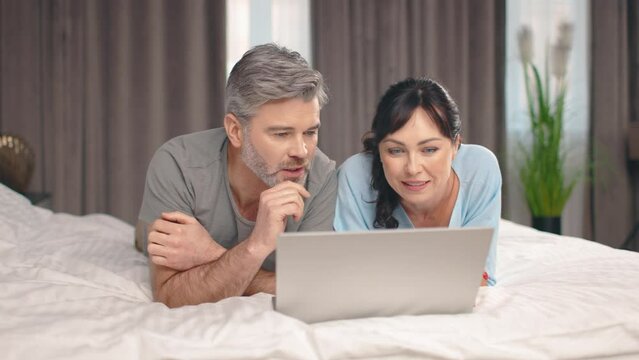Happy middle aged man and woman using laptop computer at home watching online show lying in bed together. Adult couple looking at gadget screen making video call chatting in bedroom at home.