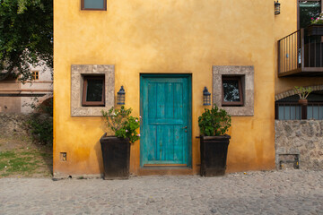 small yellow old italian style house  with a blue door in the center and two square windows at the sides and two biw flower pots in a cobbled streed - Powered by Adobe