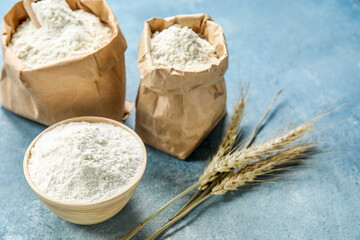 Wheat ears, paper bags and bowl with flour on blue background, closeup