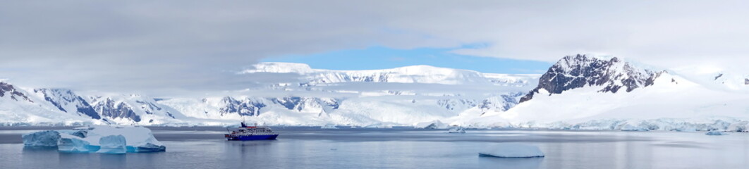 Panorama of an expedition cruise ship in a bay, surrounded by icebergs, at Portal Point, Antarctica