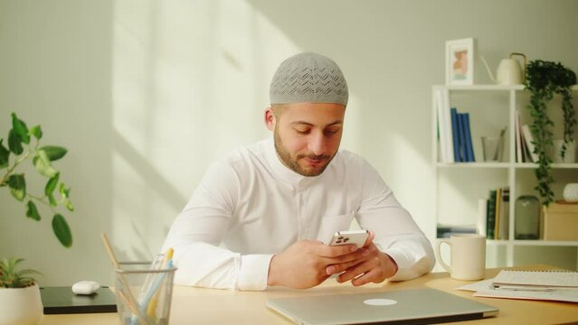 Middle eastern man using smartphone, home office. Male person texting at phone in living room. Wearing traditional Islamic clothes. Communicating with family and friends online.