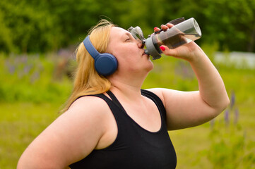 Plus size white Caucasian woman in headphones, a black T-shirt with a yoga mat in her hands is...