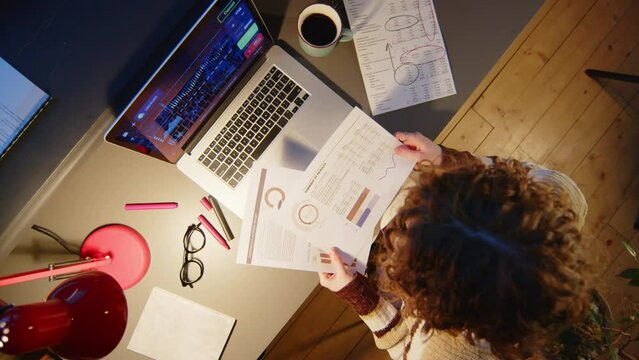 Time lapse top down camera roll shot of businesswoman using laptop, taking notes and checking financial papers at office desk while working during night