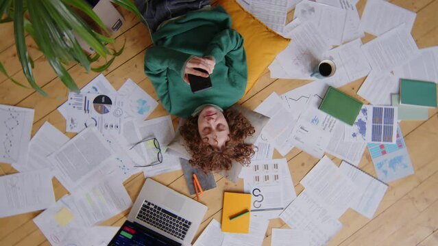 Top down camera roll shot of young female freelancer lying on floor among financial papers, texting on phone and smiling while resting during workday at home