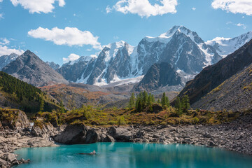 Pure turquoise alpine lake with view to forest hill and giant snow mountains in autumn sunny day. Glacial lake against huge snow covered mountain range in bright sun. Vivid colors in high mountains.