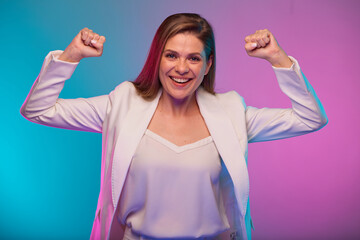 Fototapeta premium Smiling business woman bends his arms demonstrating muscles and female power, portrait with neon lights colors effect. Female model isolated on neon colored background wearing white suit.