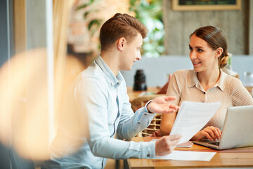 Positive young entrepreneurs in casual outfits sitting at table in coffee shop and discussing contract