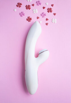 White vibrator toy for adults lies on a pink background, next to decorative hearts mimic an orgasm. Conceptual photo.