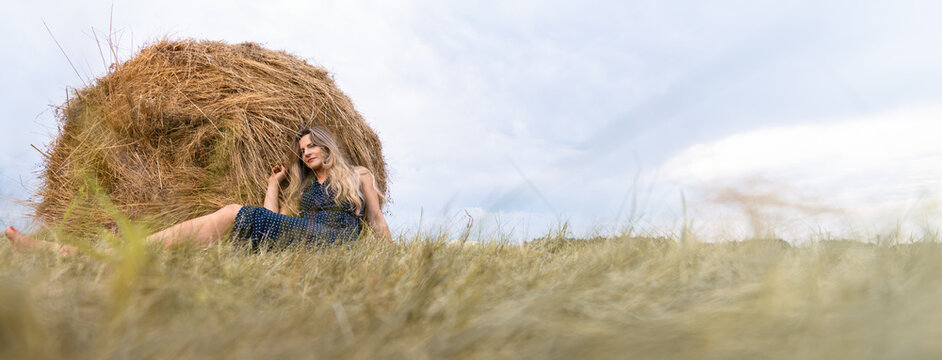 Girl and haystack. Thoughtful blonde woman in a polka-dot dress sits on the grass next to a haystack. Banner