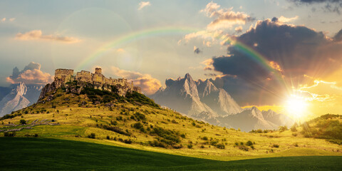 castle on the hill beneath a rainbow at sunset. composite fantasy landscape. grassy meadow in the foreground. rocky peaks of the ridge in the distant background in evening light © Pellinni