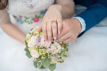 Bride wearing an elegant white dress and groom in blue suit holding hands above a delicate floral arrangement showing engagement rings and wedding bands, romantic Caucasian couple enjoying the big day