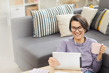 Excited young brunette woman in glasses leaning on sofa and drinking coffee while making video call via tablet app
