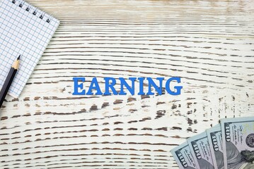 Naklejka premium EARNING - word (text) and dollars on a white wooden table, notebook, notepad. Business concept: buying, selling, commerce (copy space).