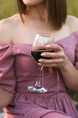 Close-up of young woman in a dress with bare shoulders holding  a glass of red wine in a field at sunset.